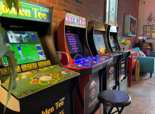 A row of classic arcade games, including Golden Tee and NBA Jam, set against a brick wall in a cozy space.