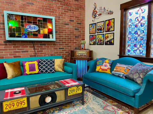 A cozy living room with teal sofas, colorful cushions, a vintage record table, and brick walls adorned with art.