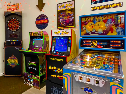 A colorful arcade room featuring classic games like Pac-Man, Teenage Mutant Ninja Turtles, and a pinball machine.