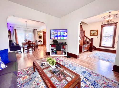 Cozy living room with a TV, coffee table, and colorful rug, leading to a dining area and hallway. Bright and inviting space.