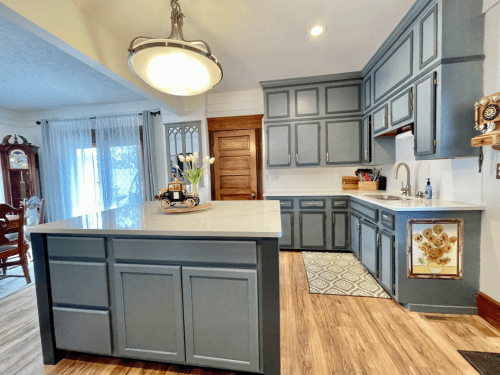 A modern kitchen with gray cabinets, a large island, and a stylish light fixture, featuring a cozy dining area in the background.