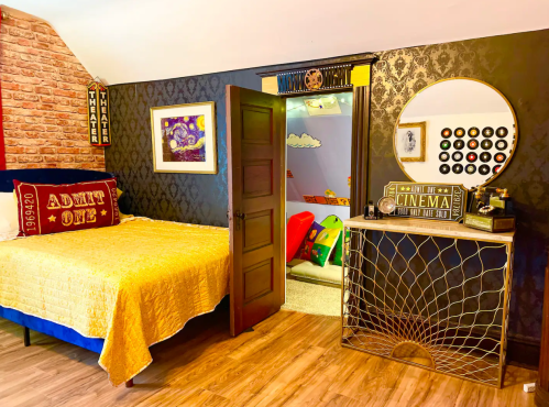Cozy bedroom with a brick wall, a gold bedspread, and a vintage cinema-themed decor featuring a round mirror and colorful play area.