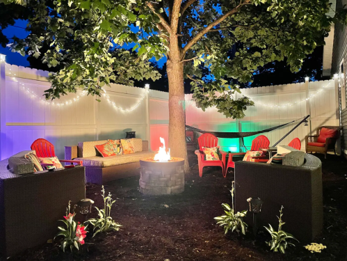 Cozy backyard setup with a fire pit, seating, and string lights under a tree, creating a warm evening atmosphere.