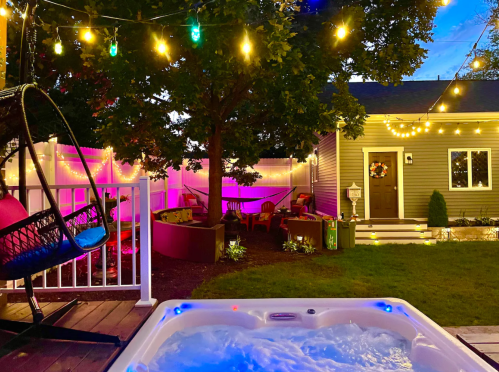 A cozy backyard scene with a hot tub, string lights, and colorful seating under a tree, creating a warm atmosphere.
