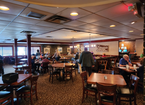 A busy restaurant interior with tables, chairs, and patrons dining and socializing. Warm lighting and decor visible.