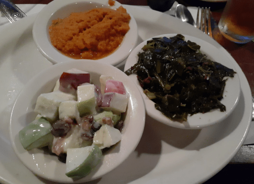 A plate with three dishes: sweet potato mash, collard greens, and a fruit salad with apples and cucumbers.