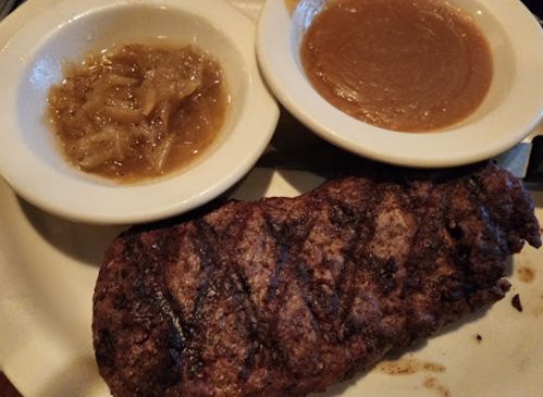 A grilled steak served with two small bowls of apple sauce and caramelized onions on a white plate.