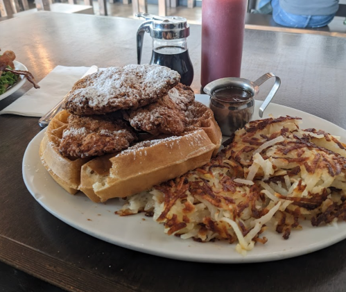A plate with a waffle topped with fried chicken, hash browns, and syrup on the side.