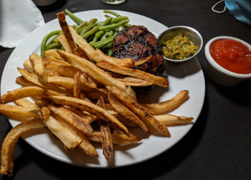 A plate with grilled steak, crispy French fries, green beans, and two sauces on the side.