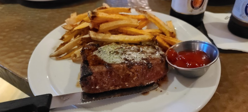 A plate with a grilled steak, crispy fries, and a small bowl of ketchup on a textured surface.