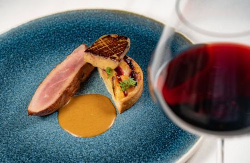 A gourmet dish featuring duck breast, seared foie gras, and a rich sauce, with a glass of red wine in the foreground.