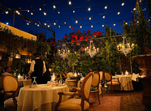 A beautifully lit outdoor restaurant with elegant tables, surrounded by greenery and twinkling lights at dusk.