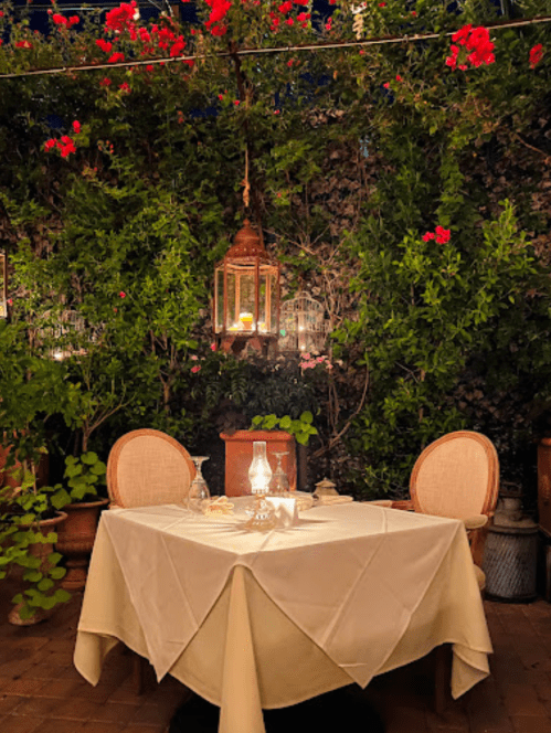 A romantic dining setup with a table, two chairs, and lanterns, surrounded by lush greenery and red flowers.