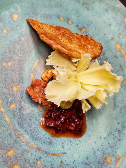 A beautifully plated dish featuring a slice of protein, garnished with a flower-like vegetable, nuts, and a sauce.