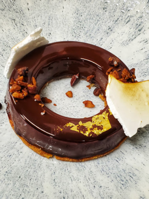 A chocolate donut-shaped dessert with a glossy glaze, topped with crushed nuts and gold leaf, on a textured plate.