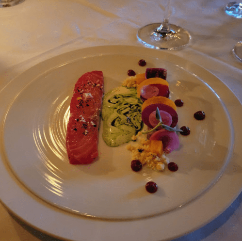 A gourmet dish featuring salmon, vibrant vegetables, and a green sauce, elegantly arranged on a white plate.