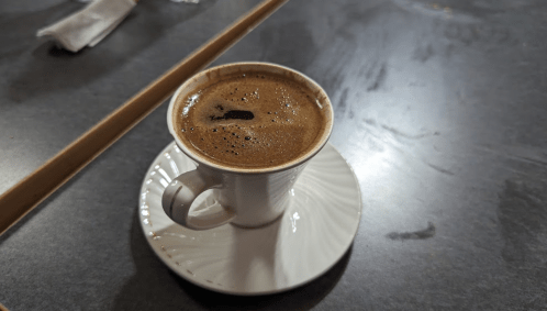 A small white coffee cup filled with dark coffee, resting on a white saucer on a dark table.