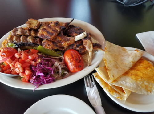 A plate of grilled meat with vegetables, including tomatoes and onions, served with warm flatbreads on the side.