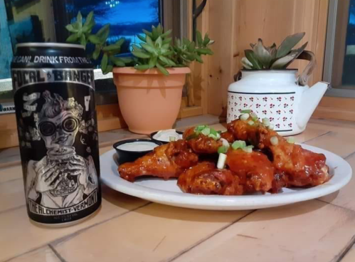 A plate of spicy chicken wings with green onions, a dipping sauce, and a can of beer on a wooden table with plants.