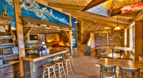 Cozy interior of a rustic bar with wooden beams, a counter, and warm lighting, featuring a mural of clouds.