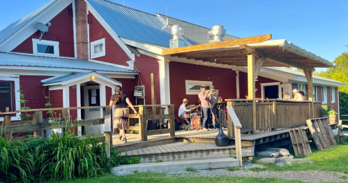 A lively outdoor scene with a band performing on a deck of a red house, surrounded by greenery and a clear blue sky.