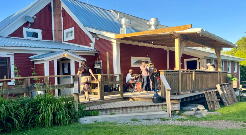 A red building with a wooden deck, where people are gathered, enjoying music and a sunny outdoor setting.
