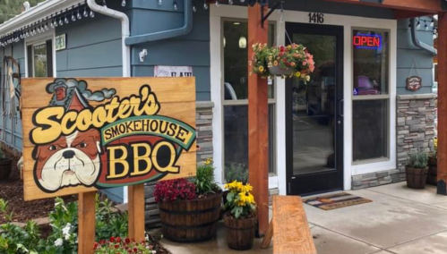 Sign for "Scooter's Smokehouse BBQ" in front of a blue building with flowers and an "Open" sign.