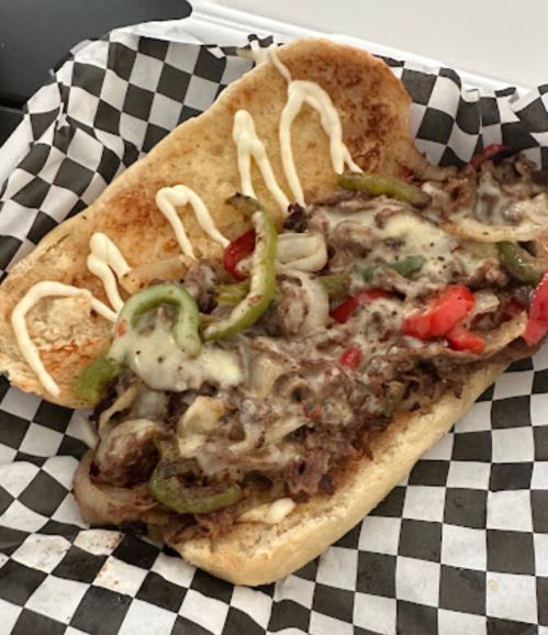 A loaded sandwich with beef, melted cheese, and peppers on a toasted bun, drizzled with mayonnaise, on a checkered paper.