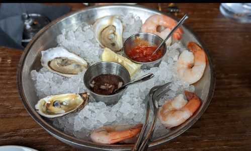A metal platter filled with ice, featuring shrimp, oysters, and dipping sauces.
