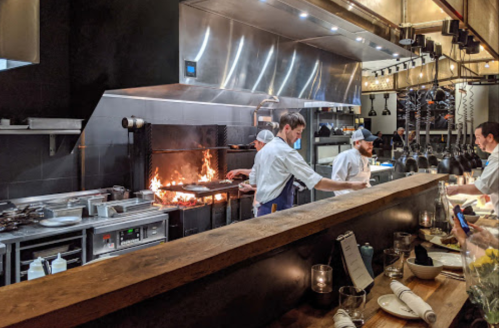 A busy restaurant kitchen with chefs cooking over an open flame, surrounded by modern equipment and a wooden counter.