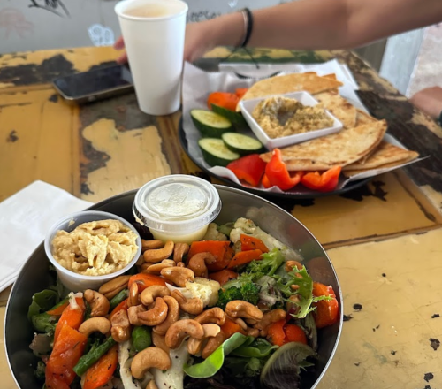 A bowl of salad topped with cashews and a side of hummus, cucumbers, and pita on a rustic table.
