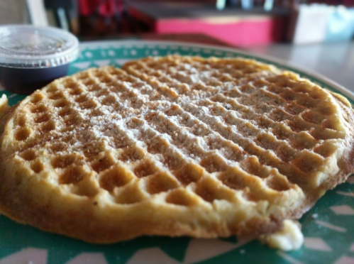 A golden waffle on a green plate, lightly dusted with powdered sugar, accompanied by a small container of syrup.