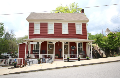 A red two-story house with white trim, a porch, and a sloped driveway, surrounded by greenery and parked cars.
