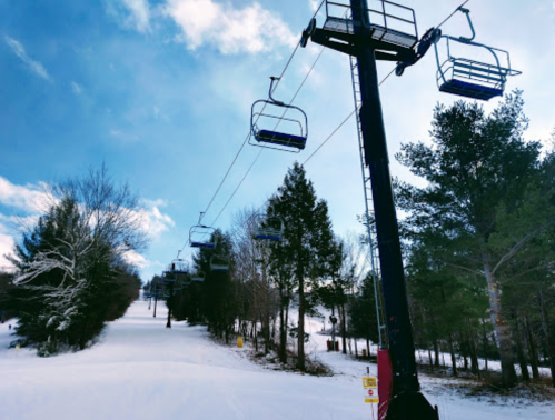 A ski lift ascends a snowy slope, surrounded by trees under a bright blue sky.