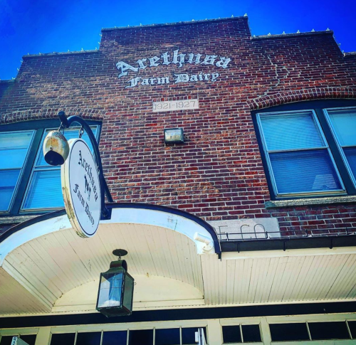 Brick building with "Arethusa Farm Dairy" sign, featuring large windows and a lantern under a blue sky.