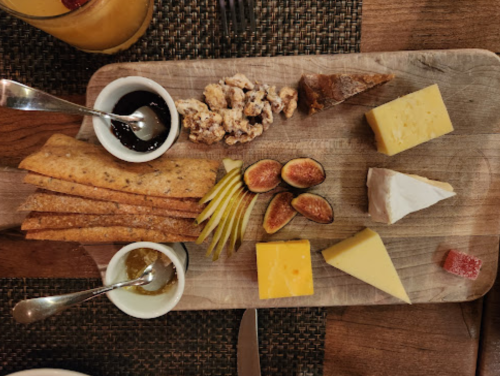 A wooden platter with assorted cheeses, figs, crackers, nuts, and small bowls of honey and jam.