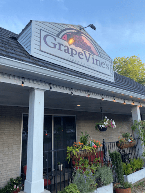 Exterior of GrapeVine's Italian Restaurant, featuring a sign, plants, and outdoor seating under a clear blue sky.
