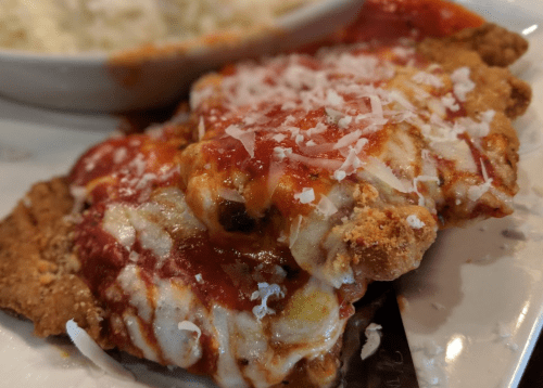 A close-up of a breaded and fried dish topped with marinara sauce and shredded cheese, served with a side of pasta.