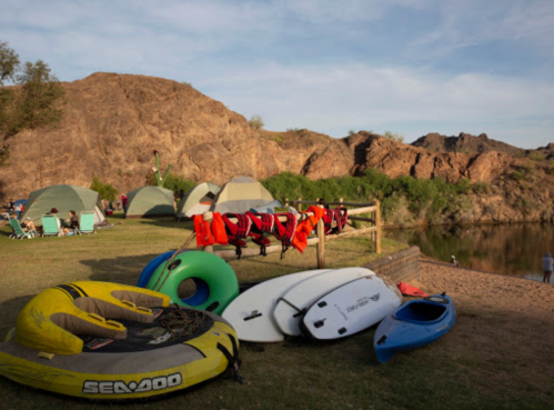 A campsite by a river with tents, kayaks, and life jackets on a rack, surrounded by rocky hills and greenery.