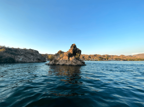 A rocky outcrop rises from calm water under a clear blue sky, surrounded by hills and greenery.