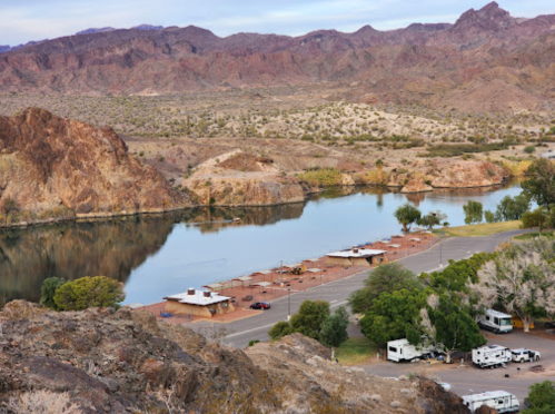 A scenic view of a river surrounded by rocky hills, with RVs parked along the shore and trees nearby.