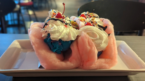 A colorful ice cream dessert topped with whipped cream, cherries, and sprinkles, served on a bed of cotton candy.
