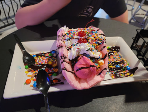 A colorful dessert featuring a pink waffle taco filled with ice cream, whipped cream, sprinkles, and a cherry on top.