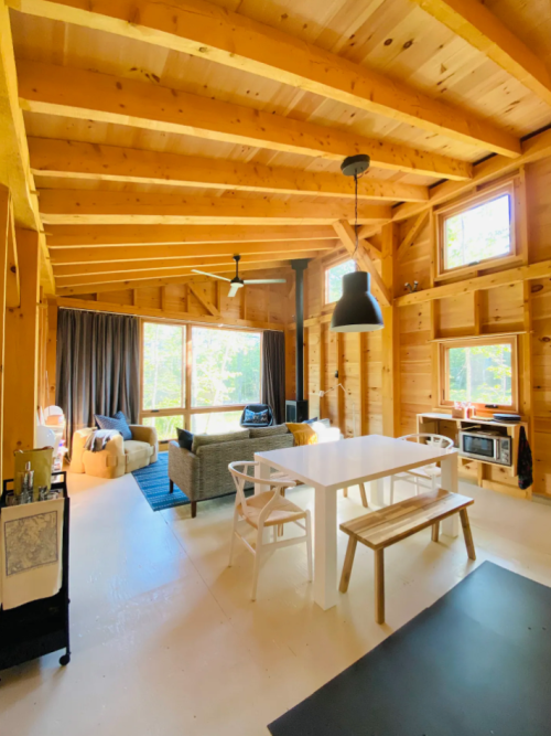 Cozy wooden interior of a cabin with a dining table, sofa, and large windows overlooking nature.
