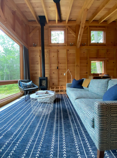 Cozy wooden living room with a patterned sofa, coffee table, and large windows overlooking greenery.