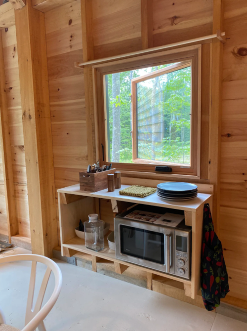 A cozy wooden interior featuring a window, microwave, and shelves with plates and utensils.