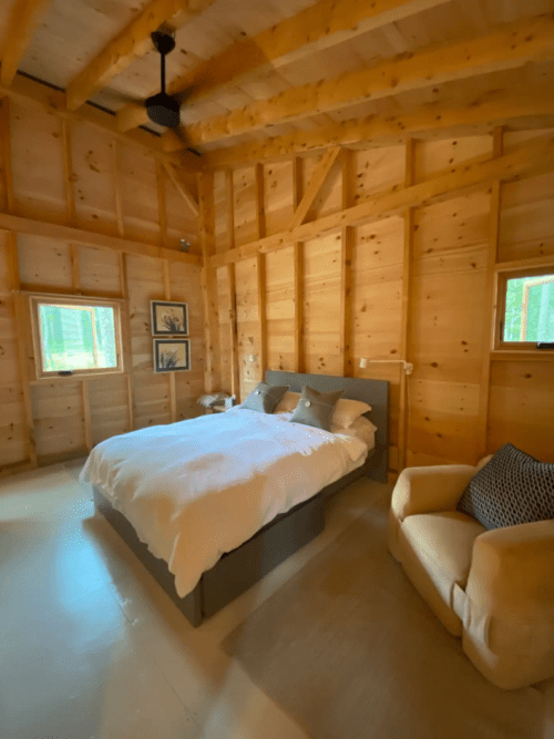 Cozy bedroom with wooden walls, a bed with white bedding, and a small armchair, surrounded by natural light.