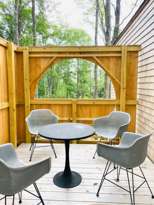 A cozy outdoor seating area with four chairs and a round table, surrounded by wooden walls and trees in the background.