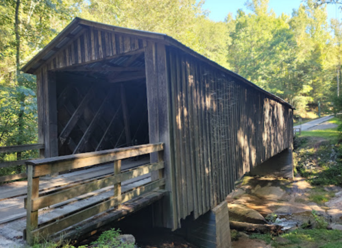 A rustic wooden covered bridge surrounded by trees, with a stone path leading to it.