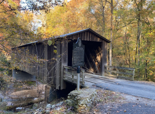 A wooden covered bridge spans a small stream, surrounded by autumn foliage and a historical marker nearby.